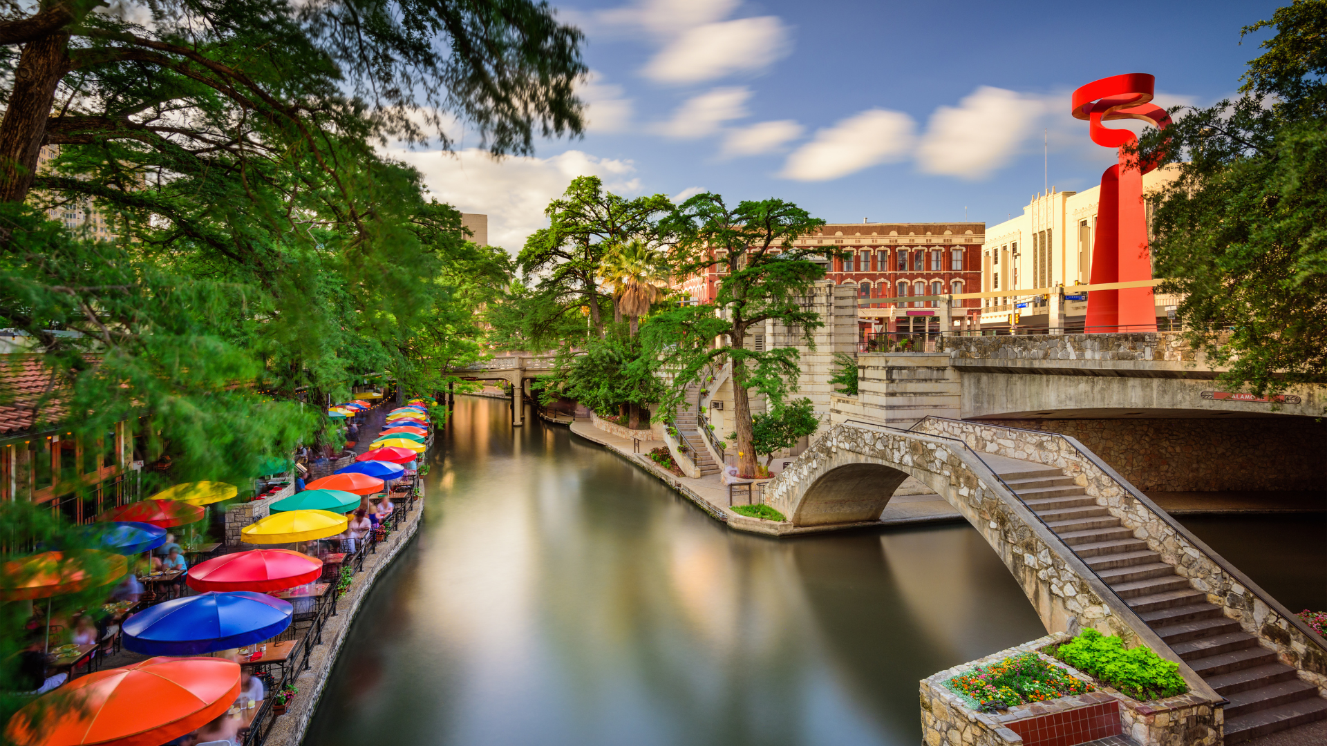 San Antonio riverwalk featuring colorful umbrellas at restaurant, trees and walking bridge